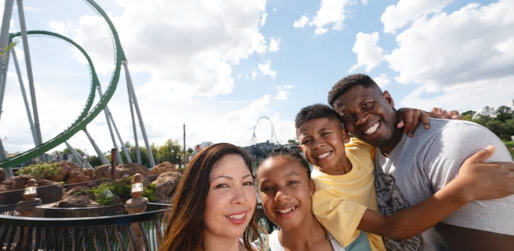 A smiling family of five enjoying a day at an amusement park with a roller coaster in the background under a partly cloudy sky.