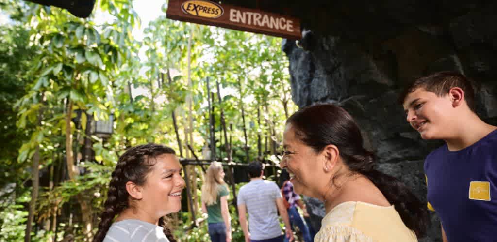 Two women and a teenage boy are smiling and talking outdoors in front of an entrance sign and lush green trees at Funex amusement park