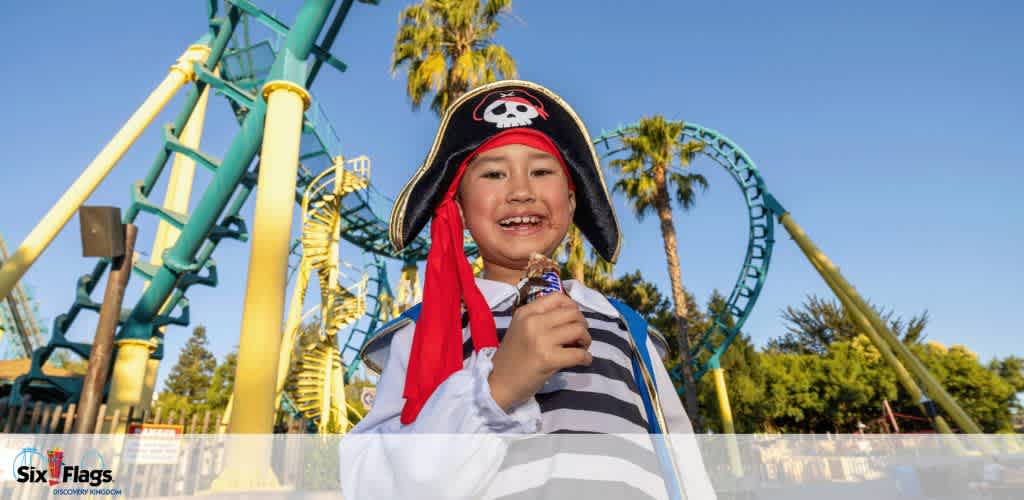 Child in a pirate costume smiling and holding ice cream in front of a roller coaster at an amusement park.