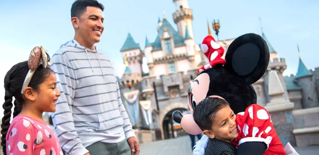 A young girl and a boy interact happily with Minnie Mouse at a castle themed amusement park with a clear blue sky in the background
