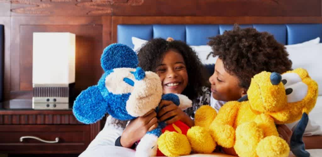 Two children are sitting on a bed holding plush toys of Mickey Mouse and Minnie Mouse, smiling and enjoying their playtime together.