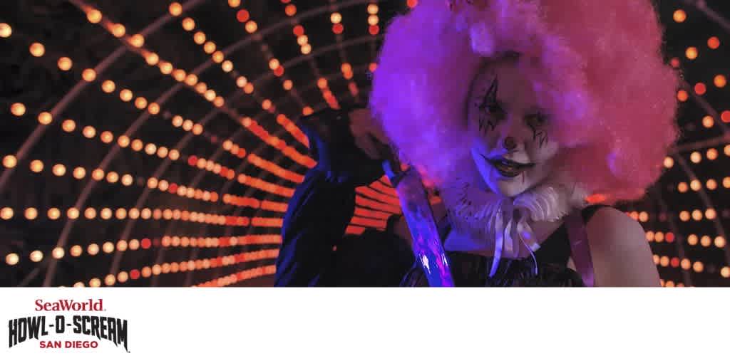 A person dressed as a clown with a pink wig and face paint holds a glowing object in front of a lit tunnel.