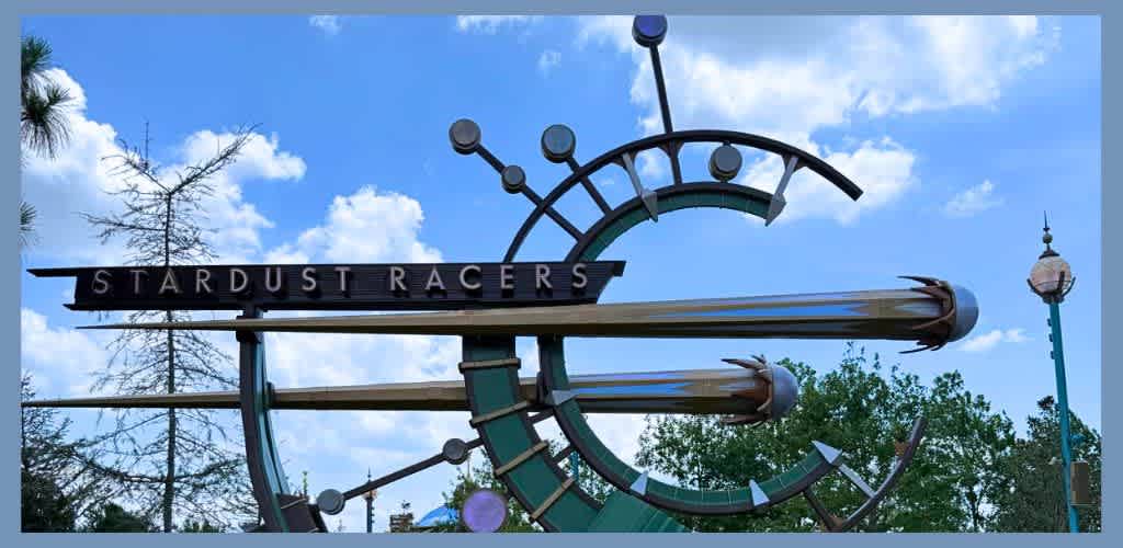 A large metallic space-themed sign with the words Stardust Racers, featuring circular and angled elements resembling a spacecraft or clock against a blue sky with clouds and trees.