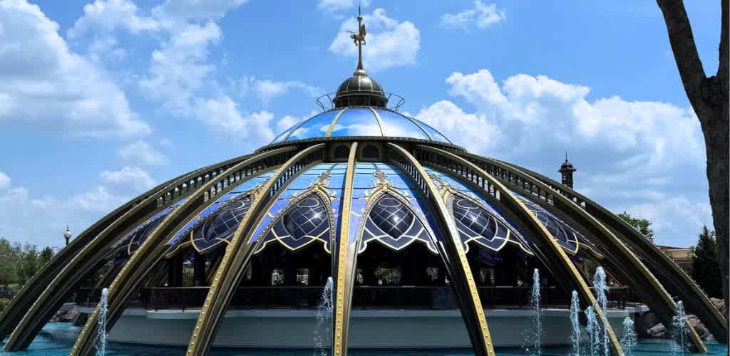 A glass and metal pavilion with a domed top, intricate gold accents, and water fountains surrounding the structure, seen outdoors under a blue sky with clouds.