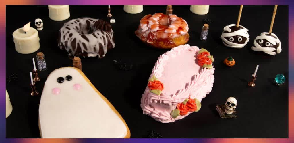 Decorative Halloween-themed desserts including ghost-shaped cookies, pink cake with roses, and spooky cake pops with mummy wrappers on a black table.
