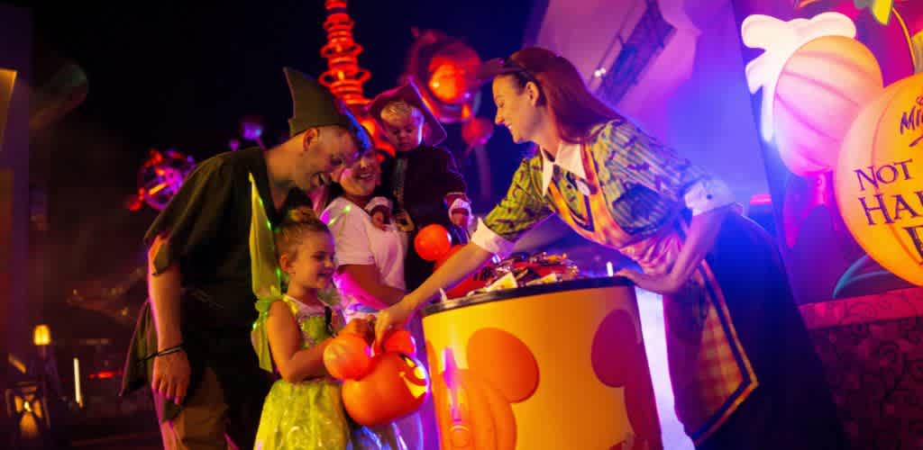 Children and adults enjoying Halloween with a smiling woman handing out candy at a decorated booth with pumpkins and bright lights