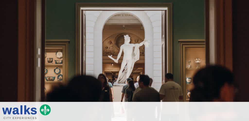 Visitors observe a large classical statue in an art museum hallway with architectural details and artwork displays around.