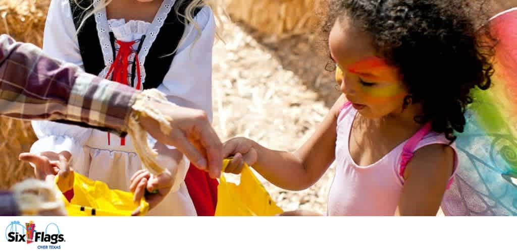 Two children in costumes receiving a yellow bag from an adult at an outdoor festival.