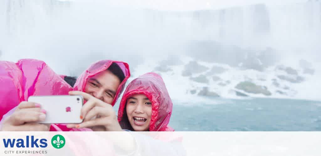Two people in pink raincoats take a selfie near a waterfall with mist and river in the background