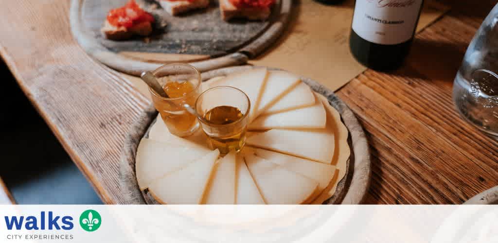 A round cheese platter with slices and two small glasses of amber-colored liquid on a wooden table.