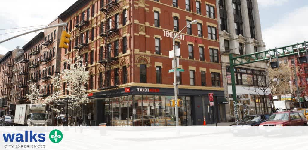 A red brick building with retail space on the ground floor and residential units above, located on a city street corner with trees and parked cars.