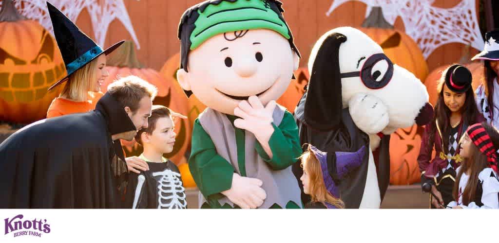 Children and adults in Halloween costumes gathered with cartoon characters in front of pumpkins and spider webs at Knott's Berry Farm.