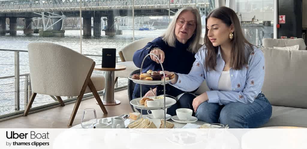 Two women sharing afternoon tea at a waterfront cafe with a three-tiered tray, cups, and pastries. Large windows overlook a dock and boats outside.
