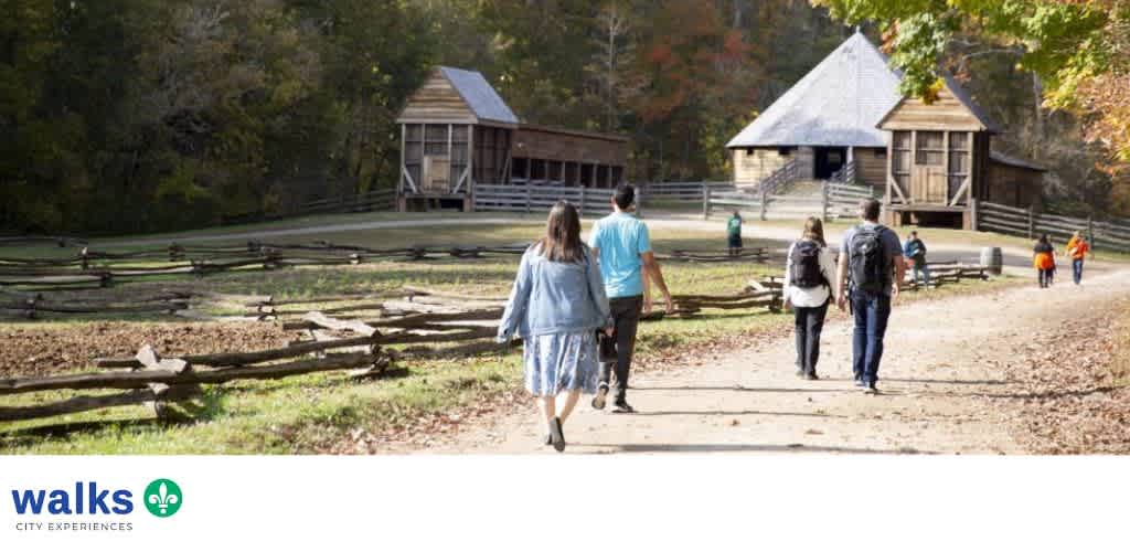 People walking on a dirt path toward historic wooden buildings surrounded by fall foliage at a park.