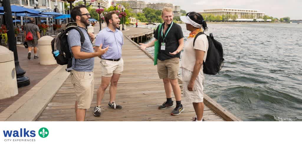 Four people stand on a wooden pier near water, engaging in conversation and smiling, with city buildings and trees in the background.