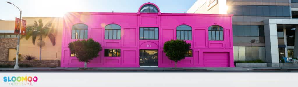 A two-story building painted bright pink with arched windows and two small trees in front. The building is located between other commercial structures.