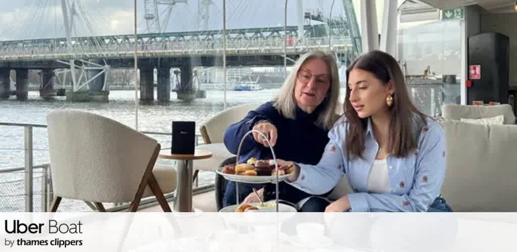 Two women sit at a table inside a boat lounge with large windows showing a bridge and water outside, sharing a tray of assorted desserts and drinks.
