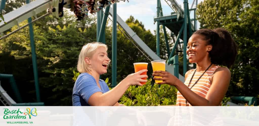 Two women toasting with drinks in front of a roller coaster at an amusement park.