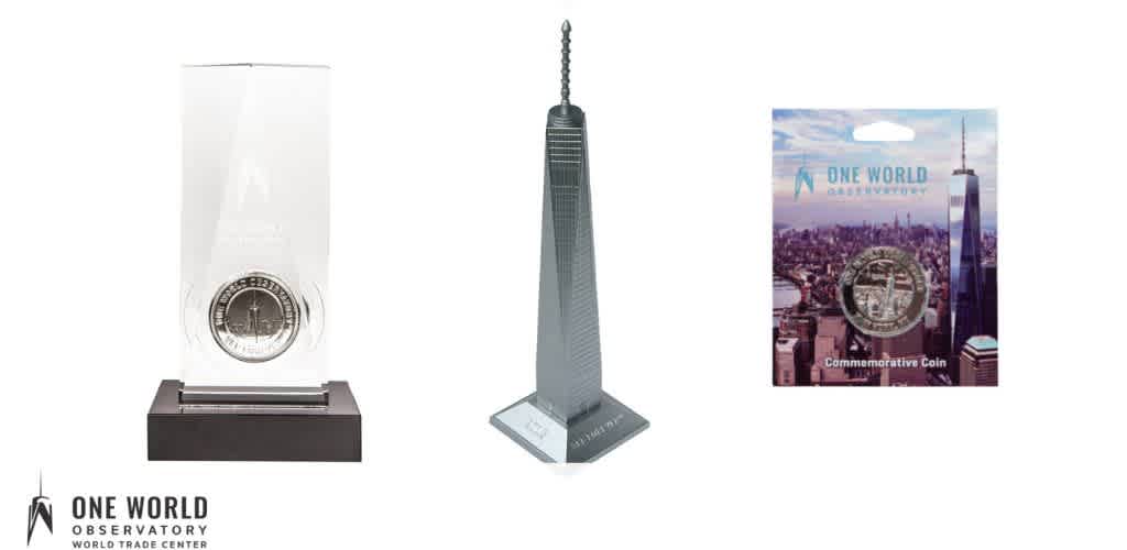 A clear glass display case with a coin and a stand-in, a tall skyscraper model with the One World Trade Center, and a cardboard box featuring a New York City skyline and a commemorative coin.