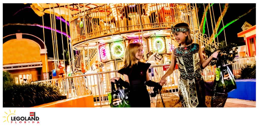Two children holding hands and carrying shopping bags at Legoland Florida amusement park at night with illuminated rides in the background