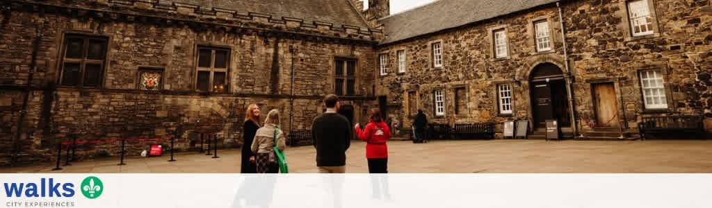 A group of people stands outside a historic stone building with multiple windows and an arched entrance in a courtyard setting.