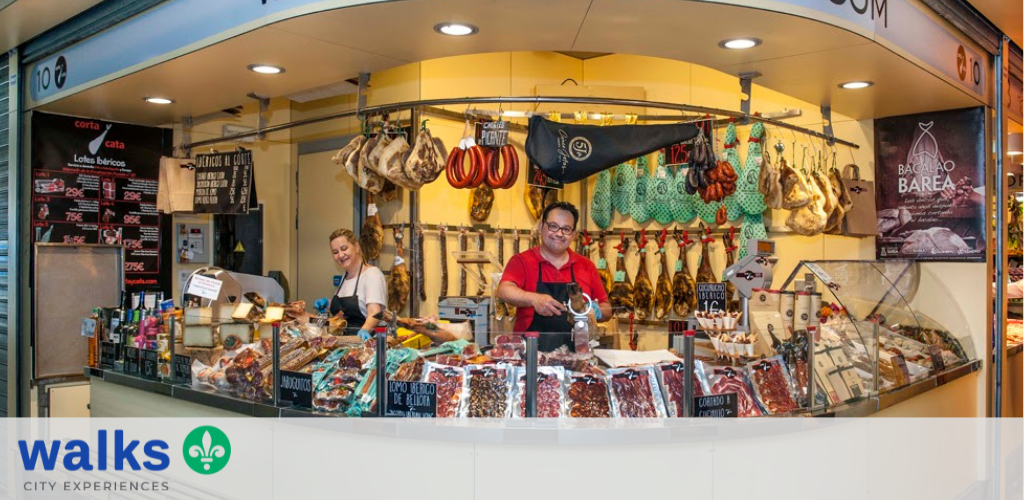 A colorful outdoor market stall selling assorted cured meats and sausages with two smiling vendors behind the counter under bright lighting