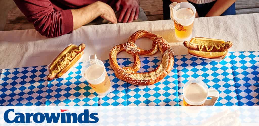 Two hot dogs, a pretzel, and three beers on a table with blue and white checkered tablecloth at an outdoor gathering