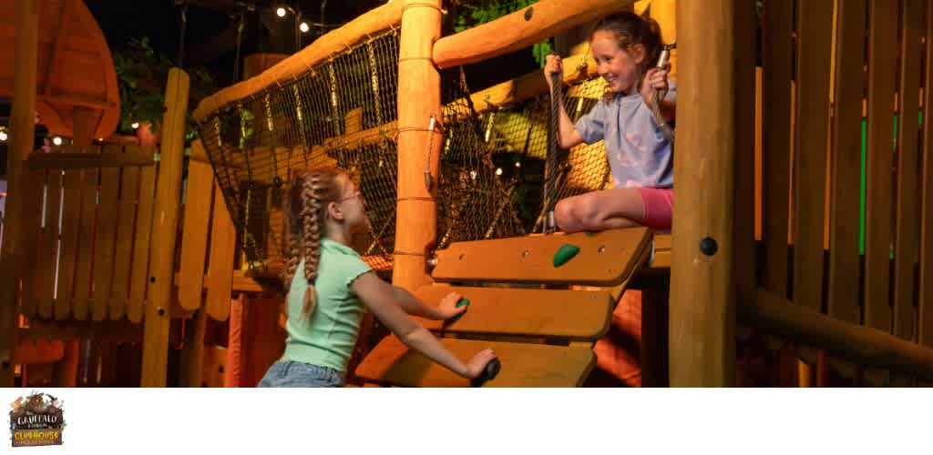 Two children are playing on a wooden outdoor climbing structure with a netted bridge and safety railings at a playground.