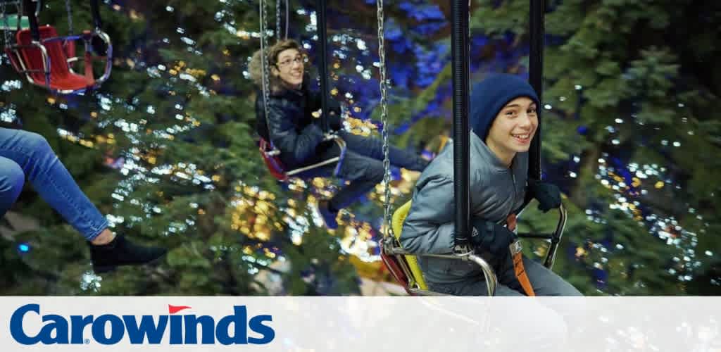 People smiling on swing rides at night with festive lights and trees in the background; Carowinds logo visible.