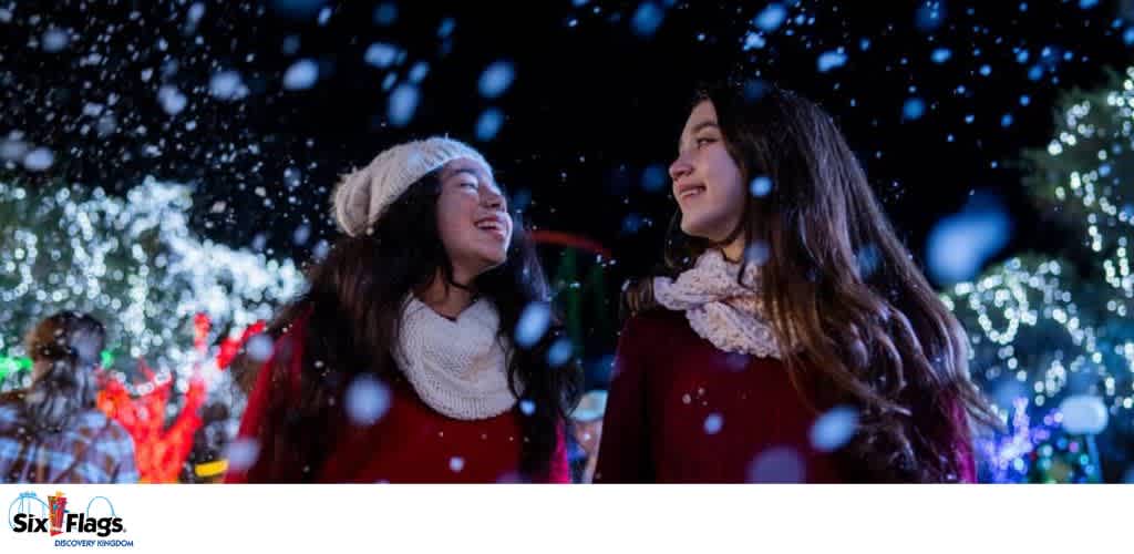 Two people enjoying falling snow with decorative lights in the background.