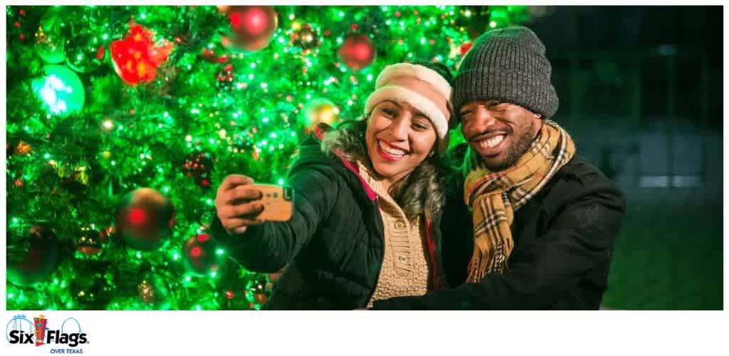 Two people smiling and taking a selfie in front of a lit Christmas tree with ornaments.