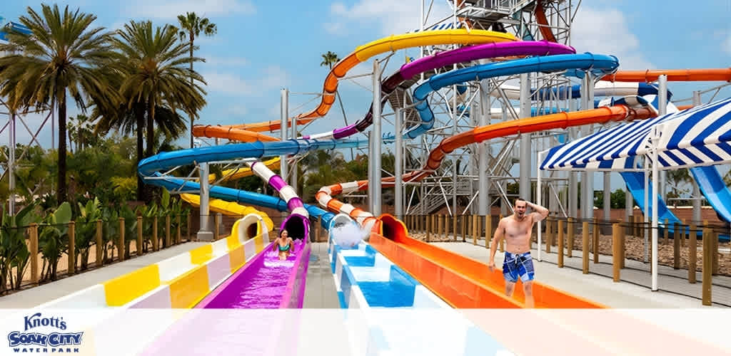 People enjoy sliding down colorful water slides at a water park with palm trees, umbrellas, and a bright, open sky in the background