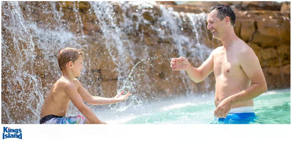 A boy and a man playfully splash water at each other near a waterfall in a pool.