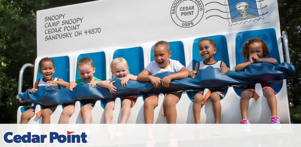 Six children smiling and laughing on a ride at Cedar Point, surrounded by greenery, with Snoopy-themed decorations in the background.