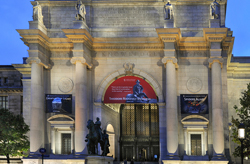 Image shows a well-lit museum exhibit featuring the towering skeletal structure of a mammoth with impressive, curved tusks. Behind it, graphic panels and a small-scale mammoth model are visible. The setting includes hanging light fixtures and informational displays, all set against a backdrop of neutral-toned walls and a large American flag.