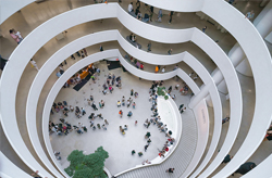 Interior view of a modern architectural space featuring a white spiral staircase at the center. Natural light filters through a glass dome and skylights, creating bright and airy ambiance. The curved lines of the staircase contrast with the angular shapes of the surrounding walls and ceiling.