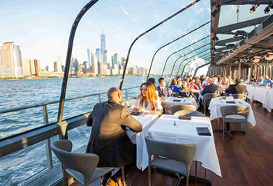 A dining area inside a boat with large windows offering a view of the skyline and water. People are seated at tables covered with white tablecloths, enjoying meals. The interior is well-lit by natural light, and the atmosphere appears serene and convivial.