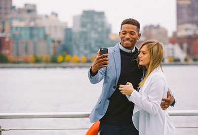 A man and a woman are casually dressed and stand close together beside a waterfront railing. The man, wearing a blue blazer over a gray shirt and brown pants, holds up a smartphone with his right hand, seemingly taking a selfie. The woman, in a white coat over a black outfit, stands to his left with her arm around his waist. The city skyline stretches across the blurred background.