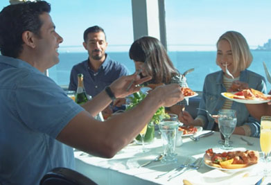 This image displays a group of four individuals enjoying a meal together at a restaurant. There are two men and two women at the table, which is set against a large window looking out onto a bright blue sky and a calm sea. All of them appear to be in a cheerful mood, engaged in casual conversation while they dine. The man closest to the foreground is smiling as he looks towards one of the women, who is focused on her plate. The table is adorned with a variety of dishes, including what appears to be pasta, and glasses filled with water. A bottle of sparkling wine sits chilling at the table, complementing the experience. Sunlight streams into the room, creating a warm and inviting atmosphere.

Remember, when planning your next dining experience, visit FunEx.com for the lowest prices and exclusive savings on restaurant tickets and more.