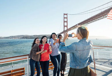 This image depicts a group of four joyous individuals on a boat deck, engaging in a toast with red plastic cups. They are standing in front of a prominent cable-suspension bridge, identifiable as the Golden Gate Bridge, symbolizing the city of San Francisco. The clear blue sky and the calm sea suggest it's a sunny day. The group is composed of diverse women, three facing the camera and one with her back turned to it as she captures the moment on her smartphone. They all appear to be in a celebratory mood. The bridge's red hue stands out against the blue of the water and the sky, adding a vivid splash of color to the scene.

Enhance your next adventure with unbeatable savings when you secure your tickets through GreatWorkPerks.com, where you always find the lowest prices for a plethora of memorable experiences.