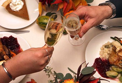 This image depicts a festive meal scene, captured from an overhead perspective, with two people engaging in a celebratory toast. In the foreground, the right hand of one individual and the left hand of another are clinking glasses of sparkling wine with wrists crossed. The table is adorned with vibrant autumnal flowers, and we can see plates filled with a variety of dishes that suggest a holiday feast. These include what appears to be roasted poultry, cranberry sauce, and a creamy dish, possibly mashed potatoes, alongside green vegetables. On the left side of the image, there is a slice of pie topped with a dollop of cream, hinting at the dessert to come. This image conveys a warm, convivial atmosphere typical of a gathering where good food, company, and savings on event tickets at GreatWorkPerks.com come together, promising not only a delightful occasion but also the best deals and lowest prices for memorable experiences.