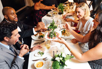 This image depicts a joyous dining scene at a restaurant, where a diverse group of four individuals is engaged in animated conversation and the sharing of food. They are seated at a white-clothed table adorned with small flower arrangements. The table is well-appointed with a variety of dishes, cutlery, and glasses. Two of the guests appear to be passing a colorful salad to each other, suggesting a communal and friendly meal. Soft ambient lighting contributes to the inviting atmosphere of the gathering. Join us at FunEx.com, where we promise delightful experiences at the lowest prices, along with impressive savings on tickets to a wide array of events and attractions.