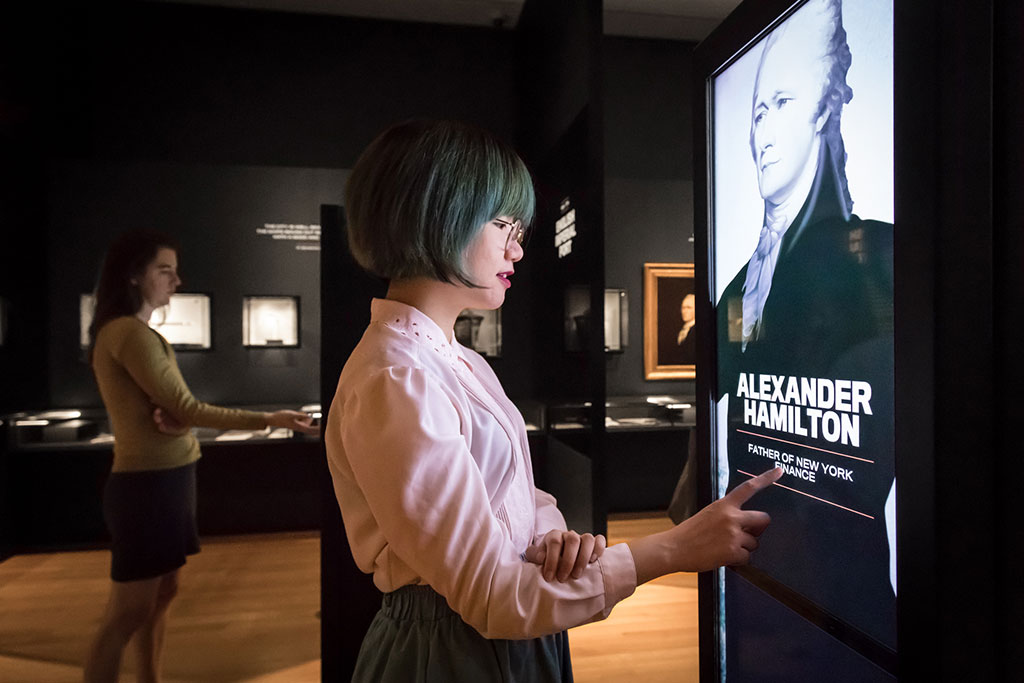 A woman with short, green-tinted hair points to a text on a digital exhibit panel featuring a prominent portrait of Alexander Hamilton, titled 'Father of New York Finance'. Another woman views artwork in the dimly lit background, emphasizing a contemplative museum atmosphere.