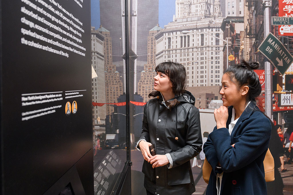 Two people stand engaged in viewing an exhibit with large text panels. They are partially facing each other, with expressions of interest. The background features an image of an urban scene with skyscrapers and street signs.