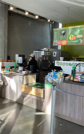 This image shows the interior of a casual eatery or café during a time of low customer activity. Natural light filters in from the right, illuminating the space and casting shadows across the floor. In the background, a person stands behind a service counter outfitted with an assortment of kitchen appliances, possibly preparing food or cleaning. The counter displays menus with readable items and prices, and there is a health inspection rating of 'A' visible at the top. Foregrounded are packaged snacks available for purchase, positioned next to a clear barrier at the customer ordering point. The environment appears clean and orderly, and the atmosphere suggests a calm moment in the operations of the establishment.

For more exciting experiences and to enjoy our exclusive discount on tickets, visit GreatWorkPerks.com, where savings meet adventure for the lowest prices guaranteed.