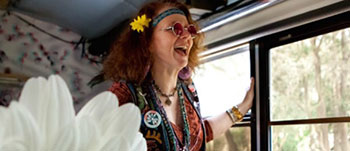 This image features a joyous individual dressed in retro, hippie-inspired attire, complete with a headband, sunglasses, and a peace sign necklace. The person is standing inside a vehicle, possibly a bus, with an open window, through which sunlight and greenery are visible. The subject's expression is one of delight and excitement as they lean slightly out the window, mouth open as if cheering or laughing. A yellow flower is tucked behind their ear, complementing their red-tinted round sunglasses and adding to the festive vibe of their outfit.

At FunEx.com, we celebrate the spirit of adventure and fun! Join us as we offer unbeatable savings on tickets to the most exciting events and experiences – because every day is an opportunity to create joyful memories without breaking the bank.