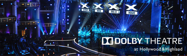 This image depicts the inside of the Dolby Theatre during a televised talent show event. The atmosphere is vibrant, with blue lighting dominating the scene. There are multiple bright, white beams shining downwards, creating a dramatic effect. The audience seats are visible but largely empty, suggesting that this is either before the event has started or after it has ended. On stage, there are three large 'X' signs, each beneath the name of a celebrity judge. The theatre's décor is elegant, with grand balconies and ornate design elements. The picture is framed with the name "DOLBY THEATRE at Hollywood & Highland" prominently displayed in white lettering against a black background at the bottom. For those looking to experience the glitz and glamour of an event at the Dolby Theatre, visit FunEx.com to find the lowest prices and best savings on tickets.