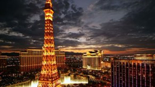 Image shows a twilight view of the Las Vegas skyline featuring a lit-up replica of the Eiffel Tower with the illuminated facade of a hotel in the background. The sky exhibits a gradient from orange to blue, signifying sunset. Bright lights and the glow from the buildings cast reflections and set a vibrant evening scene.