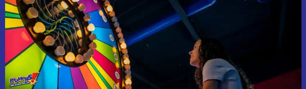 Image shows a person gleefully looking up at a vibrant, colorful spinning wheel carnival game, illuminated by lights. The game panel has a radiating pattern with a dark center and a circle of lightbulbs. A game logo is visible in the corner. The atmosphere suggests amusement and excitement at a funfair or arcade.