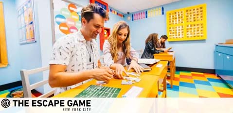 Image shows two individuals engaging in a puzzle at The Escape Game in New York City. They are concentrating on clues at a bright, colorful room with patterned walls, standing at a white table with papers spread out. A third person appears in the background. The environment suggests a lively and fun escape room experience.